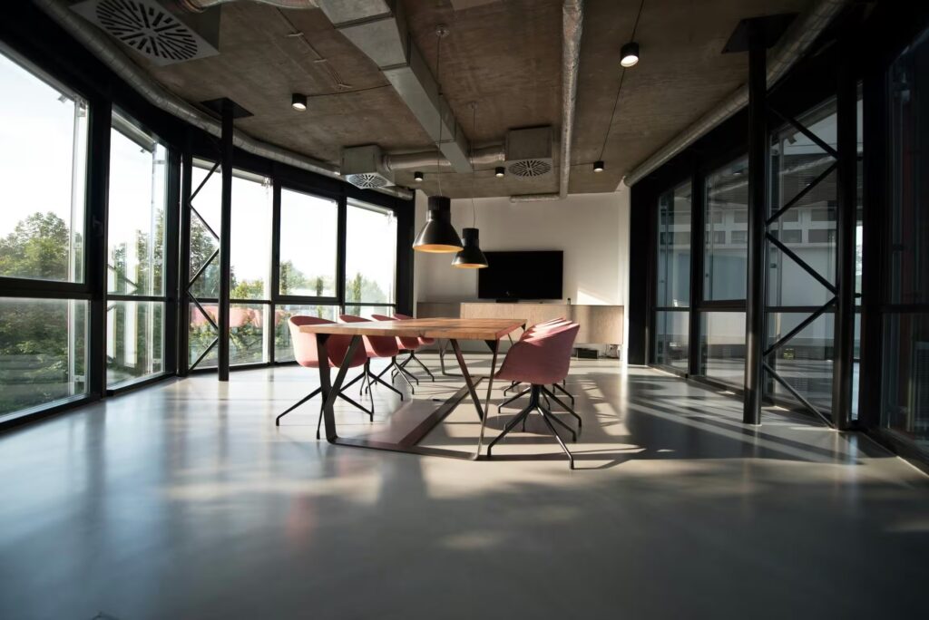 A room featuring a table surrounded by chairs, with windows allowing natural light to flow in. The decor includes elements like flooring and a ceiling, typical of indoor architecture.